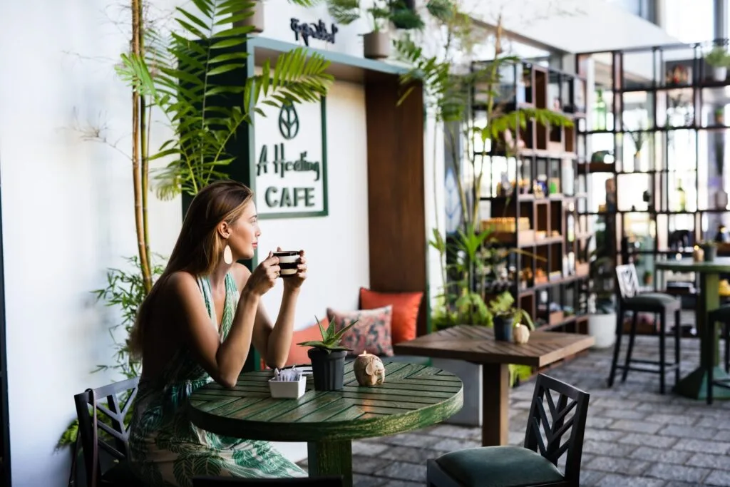 A woman enjoys a cup of coffee at a tranquil cafe with lush greenery, modern furnishings, and a stylish "Allspice Café" sign in the background, creating a serene urban oasis atmosphere.