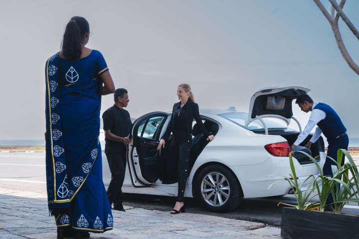 A woman in a business suit exits a white car assisted by hotel staff, with another staff member observing, all against a coastal backdrop.