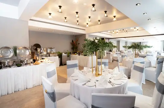 An elegant banquet hall set up for an event, with round tables draped in white linen and adorned with tall floral centerpieces. A buffet station is ready for guests at the back, under a ceiling with modern downlights.