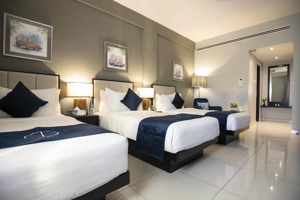 A well-appointed standard double room at Hotel Maradha with two double beds featuring white linens and dark blue throws, navy pillows, tropical-themed artwork above the beds, elegant table lamps, and a view into an en-suite bathroom reflecting the room's clean and stylish design.