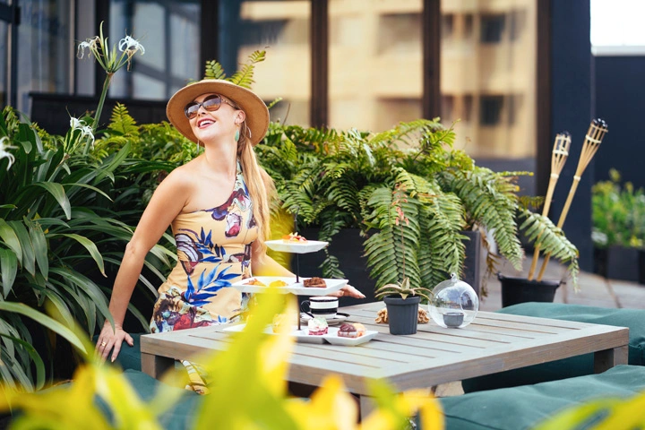 At the Healing Cafe, a joyful guest enjoys the serene rooftop ambiance, surrounded by lush greenery and the soothing presence of nature. The setting is perfect for a leisurely moment of solitude, savoring sweet treats and a refreshing beverage under the open sky.