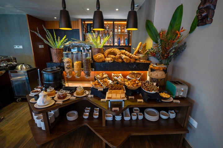 Buffet spread at Royal Code featuring an assortment of bread, cereals, and pastries, complemented by tropical greenery and modern decor.