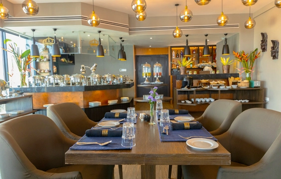 The interior of the Royal Code restaurant, featuring an elegant dining setup with brown chairs around a wooden table, ambient lighting from hanging golden lamps, and a well-stocked buffet in the background