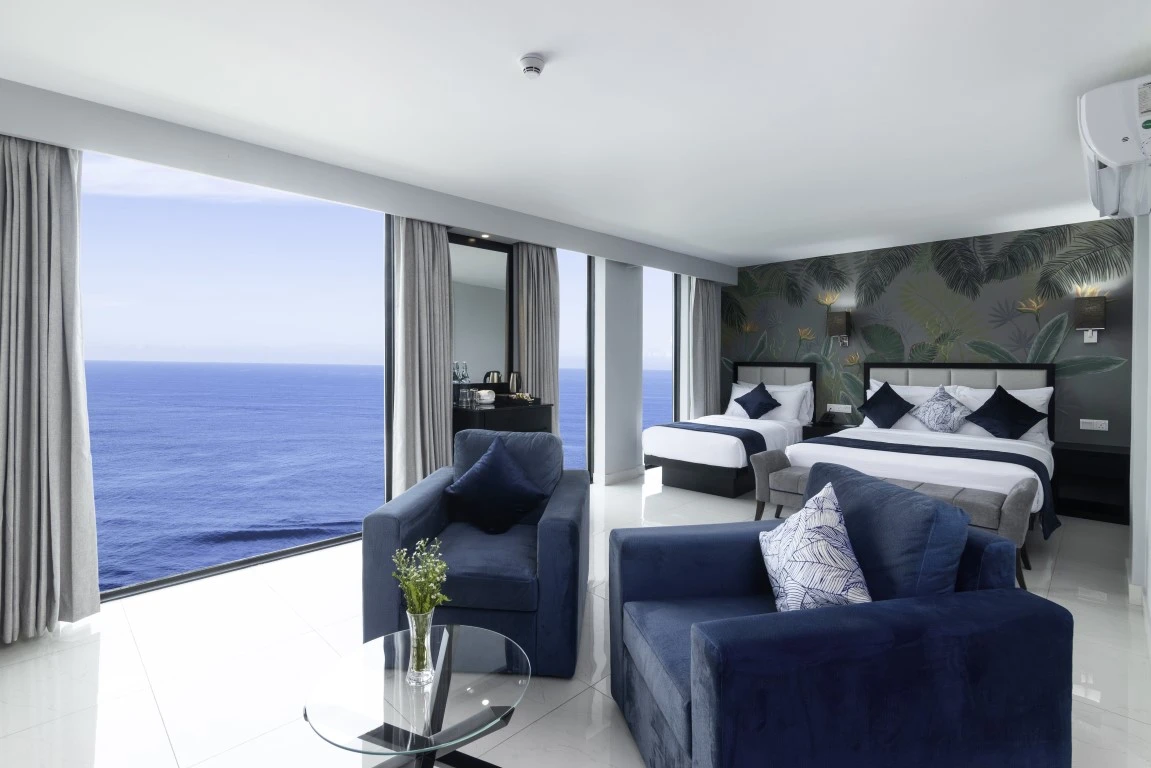 The Signature Family Sea Front room at Hotel Maradha featuring two double beds with white and navy blue bedding against a tropical-themed accent wall, comfortable blue armchairs, a glass coffee table with a bouquet, and expansive windows offering a majestic view of the sea.