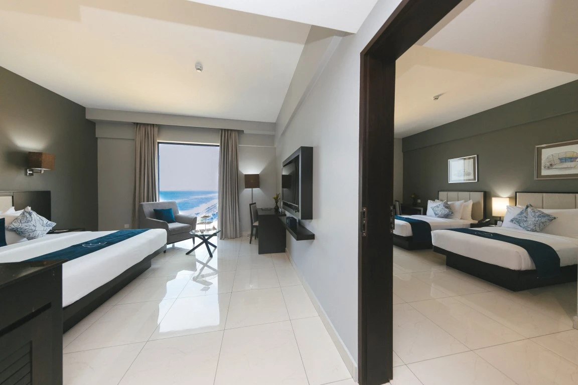 A spacious interconnected family room at Hotel Maradha with two separate bedrooms, each with a large bed dressed in white linens and navy blue runners, patterned cushions, sleek furniture, a shared sitting area, and a large window that offers a serene view of the sea.